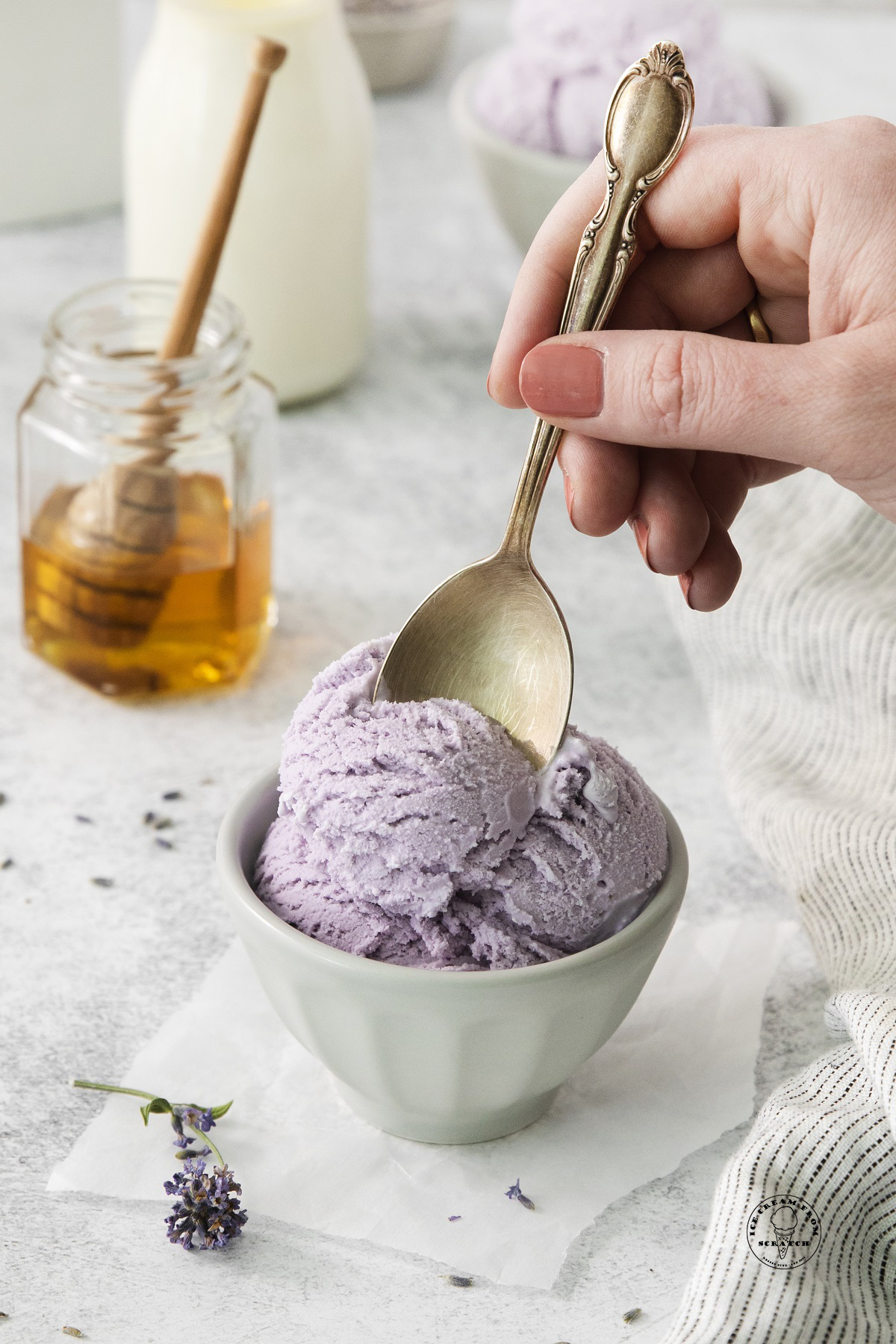 a bowl of lavender ice cream. A hand is eating it with a spoon. A jar of honey is in the background.