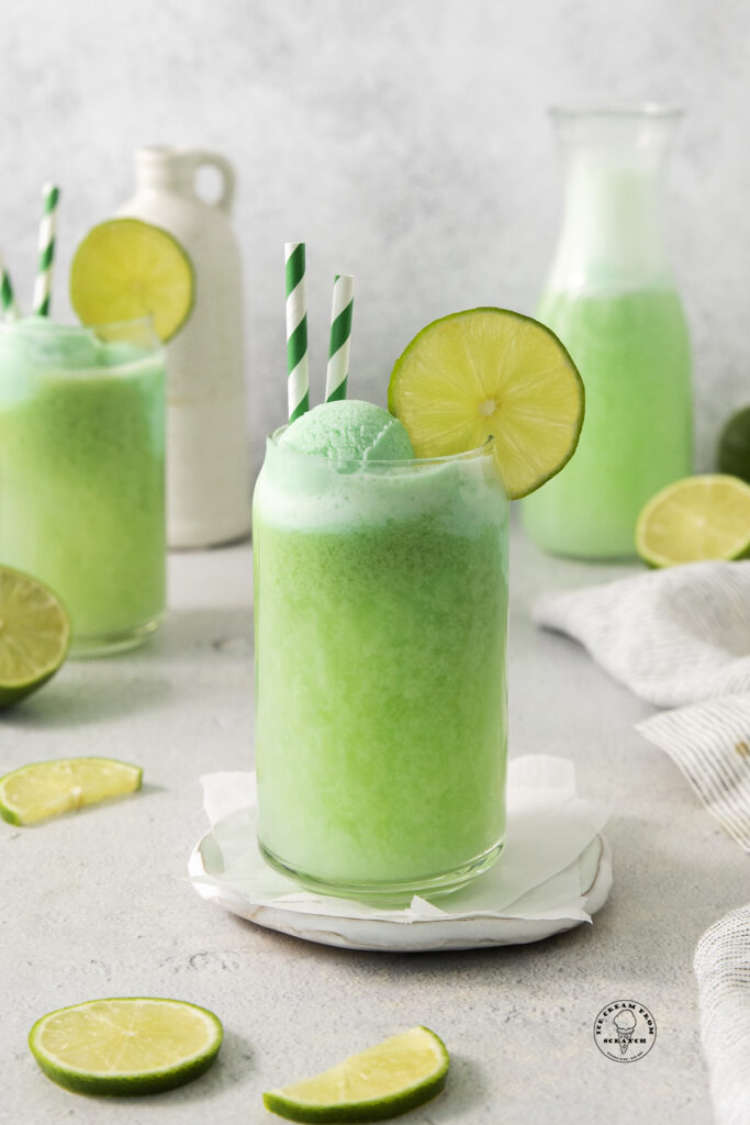 a can shaped glass filled with lime sherbet punch. It's garnished with lime wedges and two green and white straws.