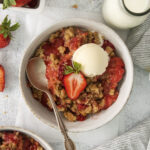 a ceramic bowl filled with warm strawberry crumble. a scoop of vanilla ice cream is melting into it.