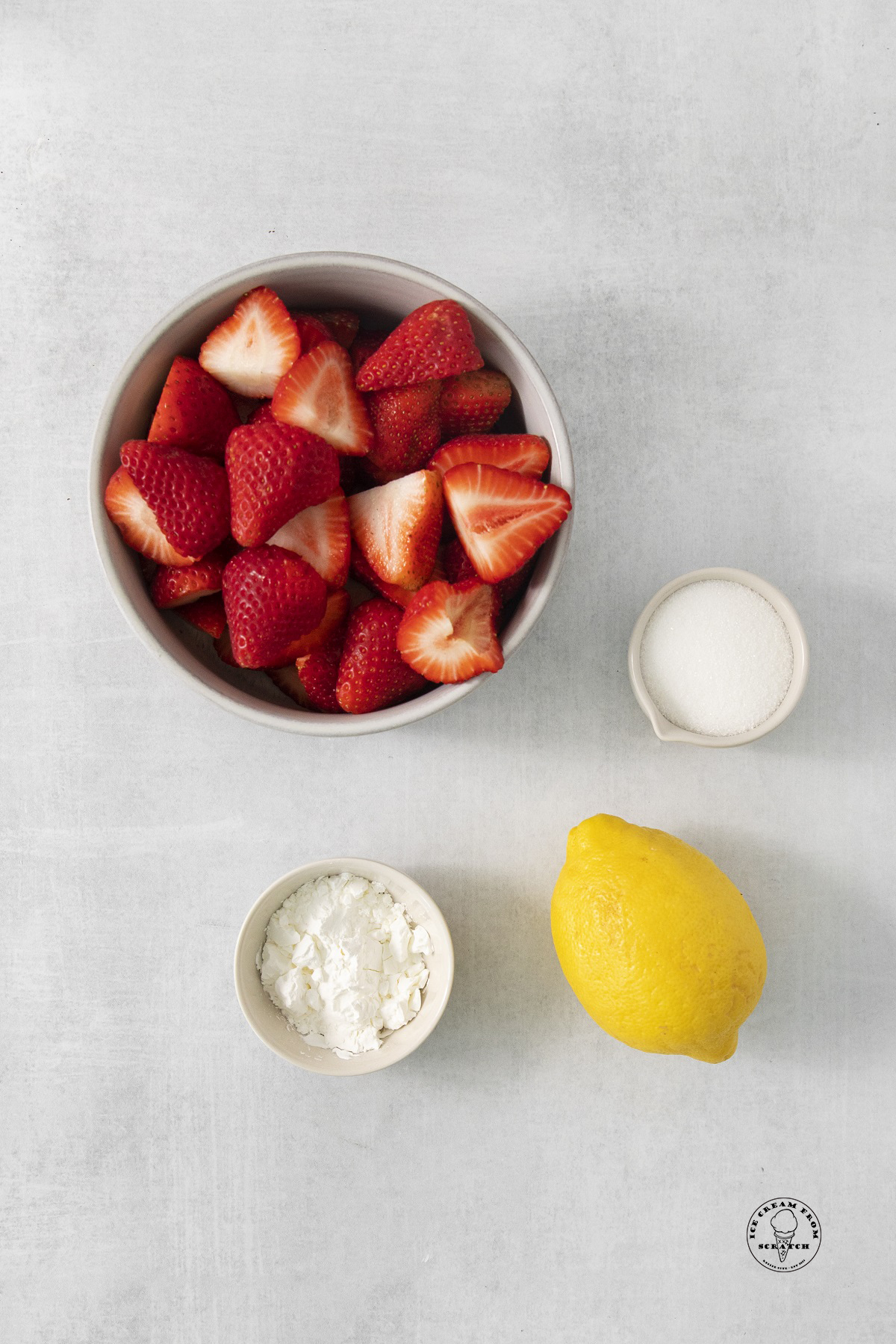 A bowl of sliced strawberries next to a lemon, a bowl of cornstarch and a bowl of sugar.