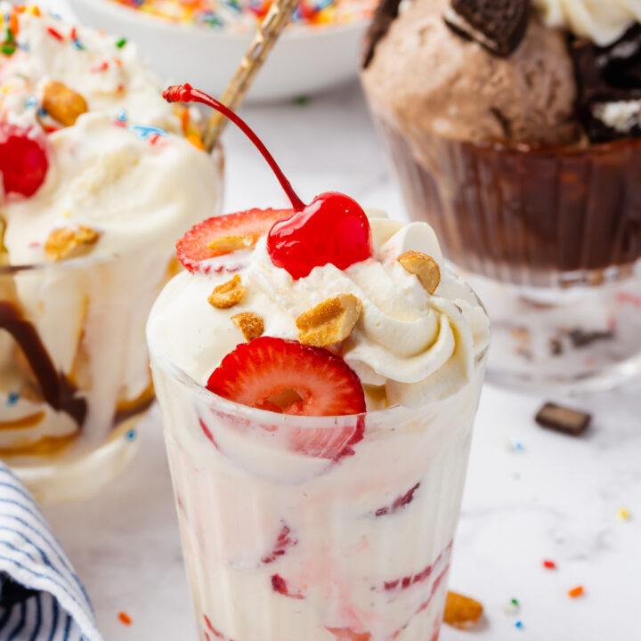 a vanilla sundae with strawberry sauce, fresh berries, whipped cream, nuts, and a cherry. Behind it are more ice cream sundaes in different styles.