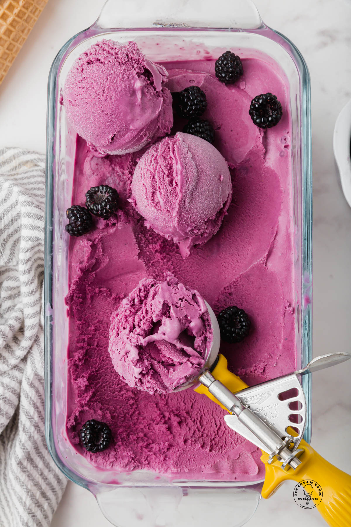a glass loaf pan of homemade blueberry ice cream, an ice cream scoop is serving it, and fresh blackberries are sprinkled over.