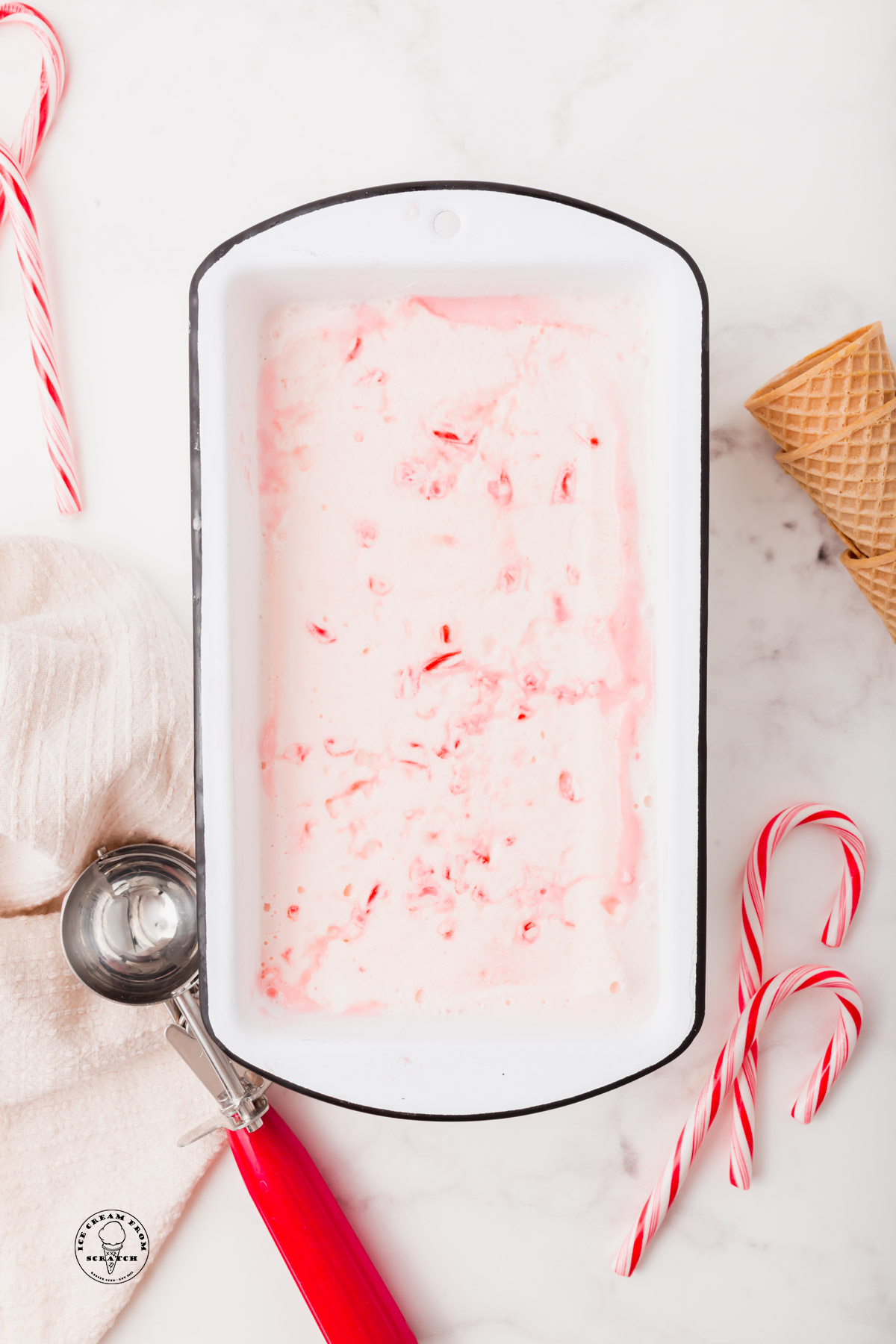 Homemade candy cane ice cream in a white bread loaf pan. Next to the pan is a red ice cream scoop, whole candy canes, and sugar cones.
