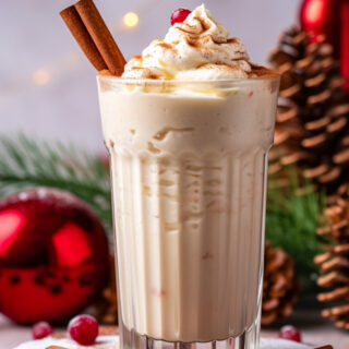 An eggnog milkshake in a tall glass, garnished with cinnamon sticks, whipped cream, and a cranberry. In the background are red ornaments, greenery, and pinecones.
