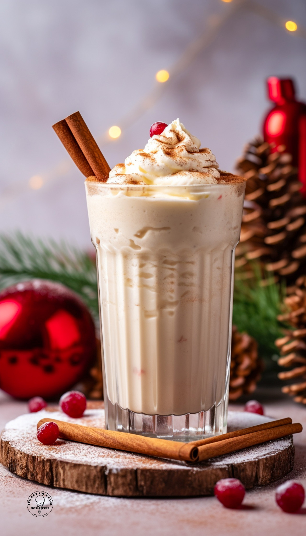 An eggnog milkshake in a tall glass, garnished with cinnamon sticks, whipped cream, and a cranberry. In the background are red ornaments, greenery, and pinecones.