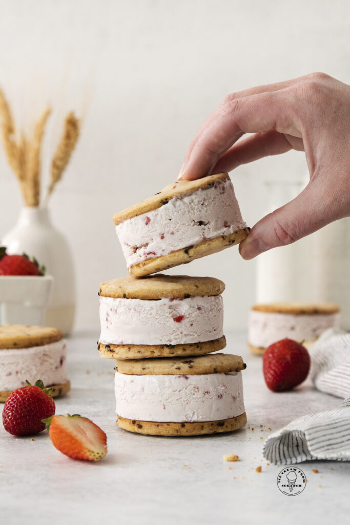 three round strawberry cookie ice cream sandwiches in a stack. A hand is picking one up.