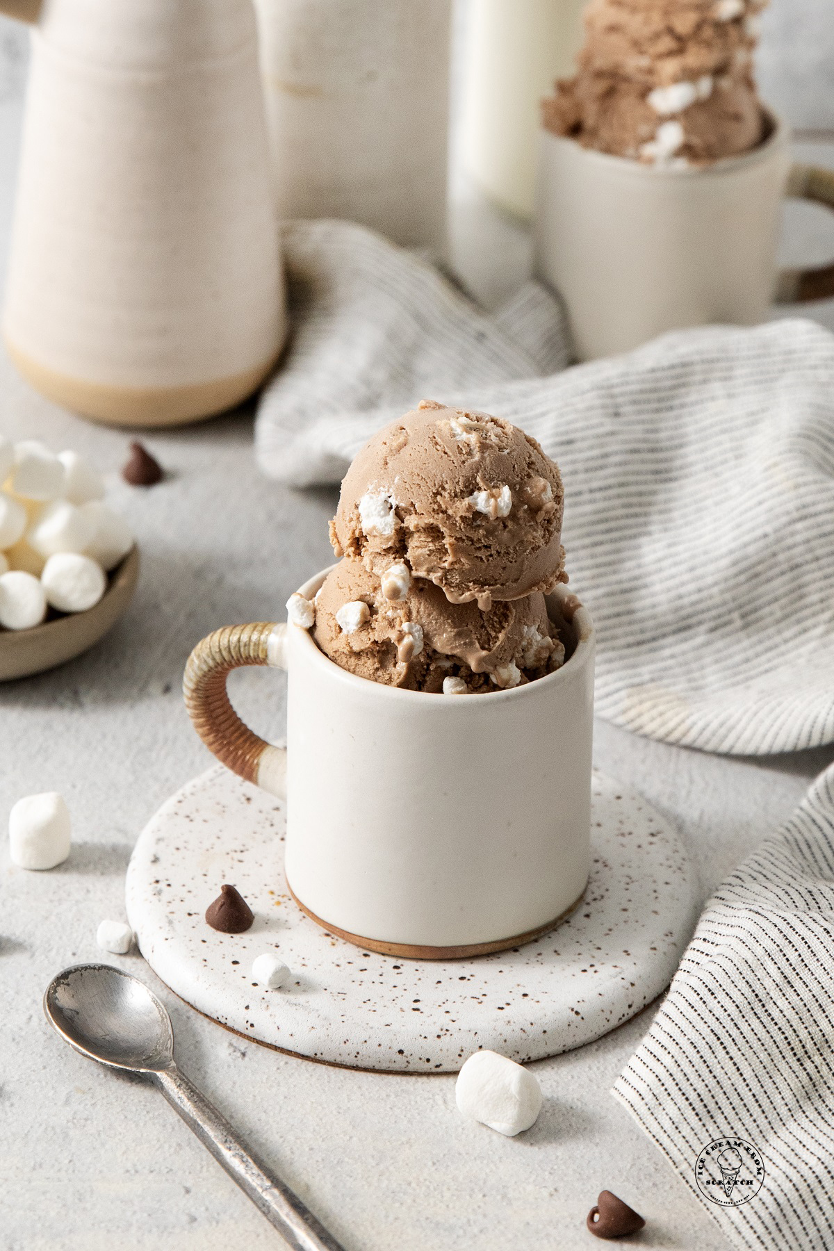 a whiter ceramic mug with a brown handle sitting on a speckled brown and white coaster, holding scooops of homemade hot chocolate ice cream. Marshmallows, chocolate chips and a small spoon are on the counter.
