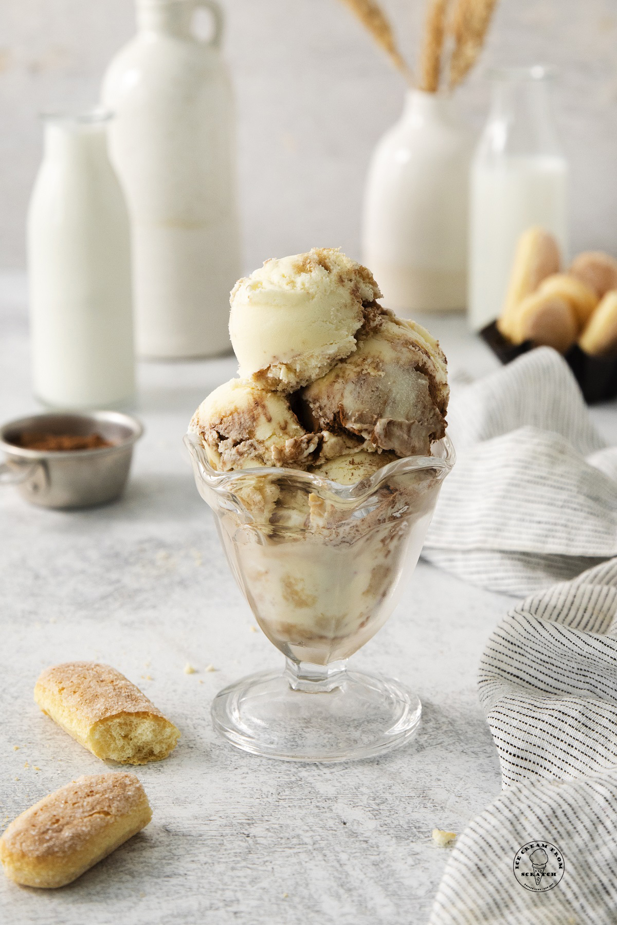 a footed ice cream dish holding scoops of homemade tiramisu ice cream. In the background are bottles of cream and ladyfingers